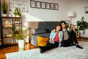 A young couple sitting on the floor of their rented apartment living room, leaning against a sofa surrounded by their belongings